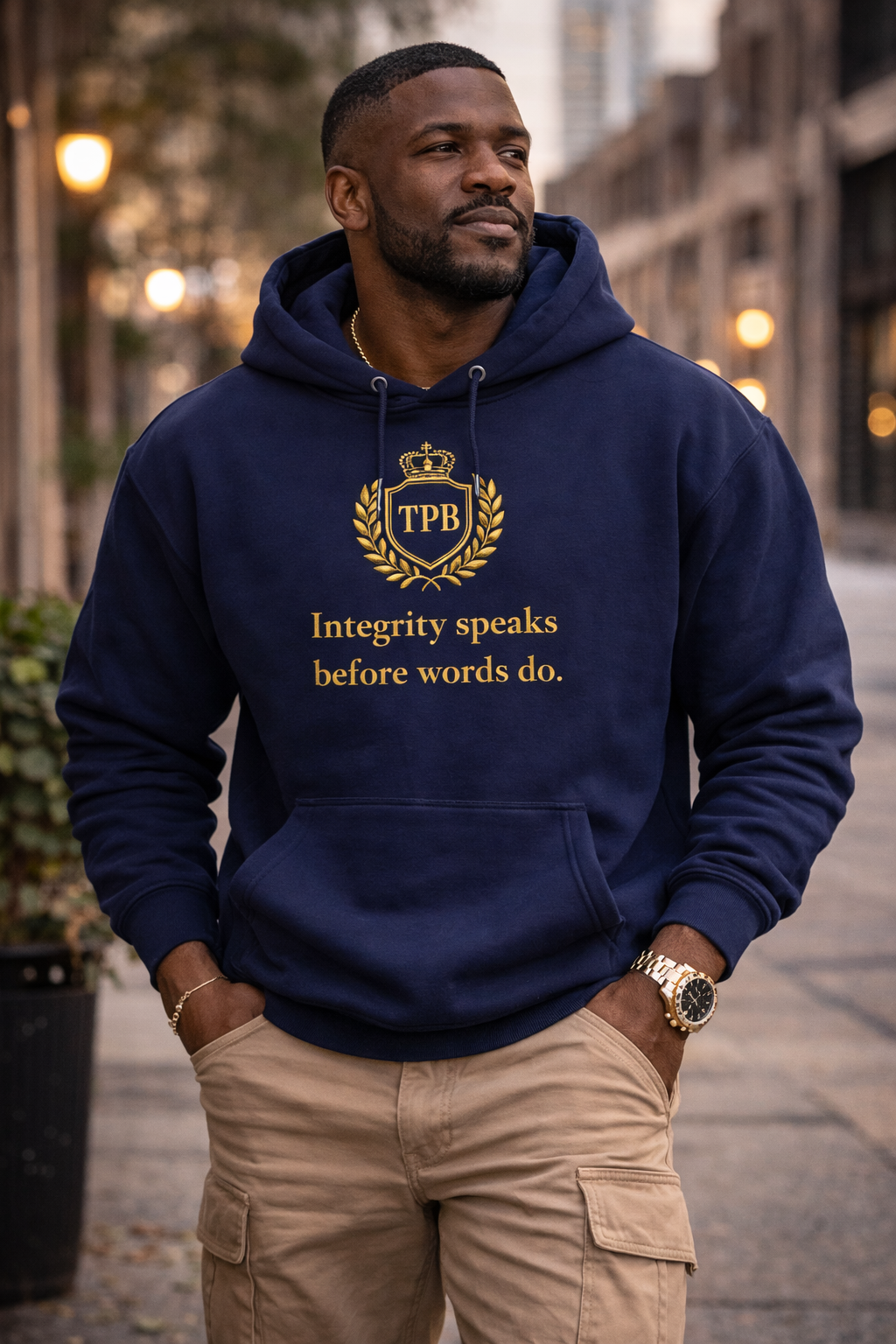 Man wearing a navy hoodie with gold text, Integrity speaks before words do, and logo, standing on a city street.